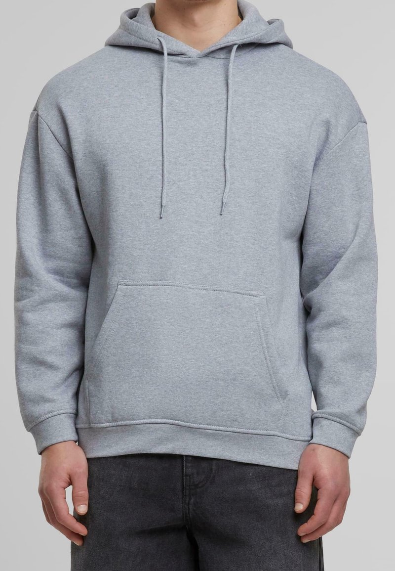 Personne portant un sweat à capuche gris clair uni avec une poche à l'avant et des cordons, associé à un pantalon foncé.