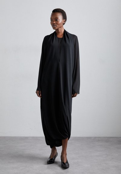 Black long-sleeve dress with a high neckline, draped silhouette, and gathered hem. Smooth fabric and minimalistic design. Worn with glossy black heels.
