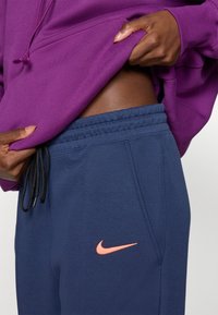 Hoodie violet avec cordon de serrage, associé à un pantalon de jogging bleu marine avec un logo Nike orange. Texture de tissu douce, détail de taille élastique.