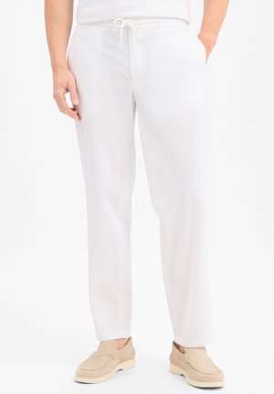 Mężczyzna w białych spodniach ze sznurkiem, ręce w kieszeniach i beżowe buty wsuwane na tle jednolitego tła.