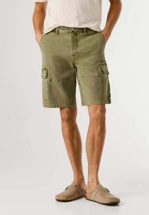 Uomo che indossa pantaloncini cargo verde oliva e scarpe beige senza lacci, con le mani in tasca, in piedi davanti a uno sfondo semplice.