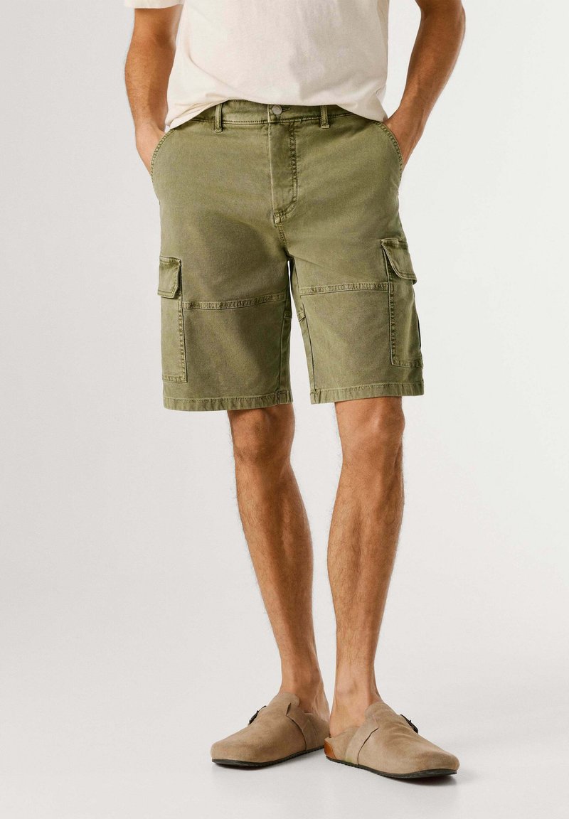 Man wearing olive green cargo shorts and beige slip-on shoes, hands in pockets, standing against a plain background.