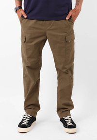 Pantalons cargo verts olive avec deux poches latérales, taille élastique et coupe décontractée, associés à des baskets noires avec des lacets blancs.