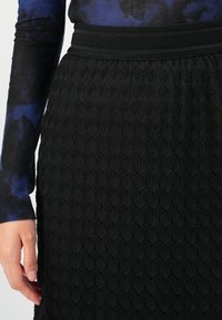 Roka, položena ob črno krilo z teksturirano diamanto in cvetličnim čipkastim vzorcem, ki je kombinirano z dolgo rokavom v temno modri in črni barvi.