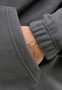 Sweatshirt en polaire gris avec tissu texturé. Doté d'une poche kangourou et de poignets côtelés. Un tatouage visible au poignet dépasse de la poche.