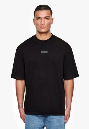 T-shirt en coton noir à col rond et manches courtes, avec une étiquette rectangulaire portant le mot "Drops" sur l'avant.