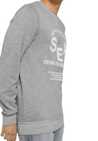 Šedá mikina s kulatým výstřihem, žebrovanými manžetami a lemem. Obsahuje potisk a texturovanou látku. Design je ležérní a volný.