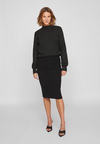 Zwart gebreide sweater met een hoge halslijn en geribbelde manchetten, gecombineerd met een bijpassende geribbelde kokerrok. Draagt zwart open teen hakken.