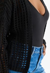 Black knitted cardigan with an open weave design, worn over a black bodysuit and high-waisted blue denim jeans. Note the textured details.