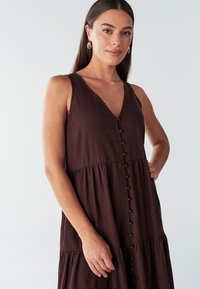 Brun ærmeløs kjole med V-udskæring, knappedetaljer ned foran og et lagdelt nederdel. Fremstillet af let stof med en subtil tekstur.