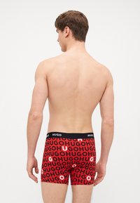 Shorts de boxer rouges avec un logo "HUGO" répétitif en noir et blanc, doté d'une ceinture élastique noire. Tissu lisse et design ajusté.