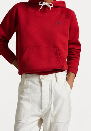 Personne portant un sweat à capuche rouge avec des cordons blancs et un petit logo, associée à un pantalon blanc taille haute, les mains dans les poches.
