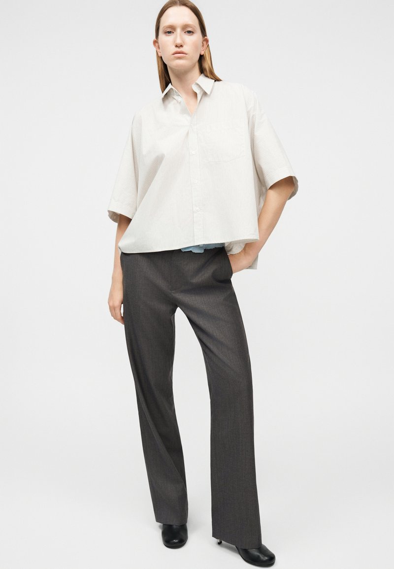 Vrouw die een losse lichtbeige overhemd met korte mouwen draagt, donkergrijze wijde broekspijpen en zwarte schoenen met hakken, staand met één hand in de zak.