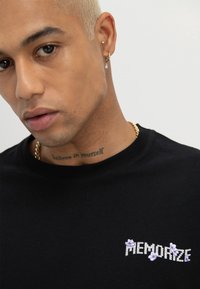 Junge Person mit kurzen blonden Haaren, Ohrringen, Hals-Tattoo mit der Aufschrift "Glaub an dich selbst", schwarzem Shirt mit dem Schriftzug "MEMORIZE" und einem violetten Blumendesign.