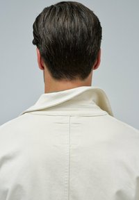 Weiße Jacke mit weicher Textur, die einen leicht erhöhten Kragen und saubere Nähte aufweist. Der Rücken zeigt minimale Akzentdetails und ein einfaches Design.
