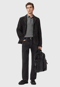 Ung mand iført sort ruskindsjakke, grå polo T-shirt, sorte jeans og sorte sko, holder en sort læder rygsæk ved sin side.