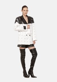 Dwurzędowa marynarka z białej tkaniny z czarnymi logo, kontrastującymi czarnymi skórzanymi klapami i dobrze dopasowanym krojem. Połączona z czarnymi szortami i wysokimi kozakami za kolano.