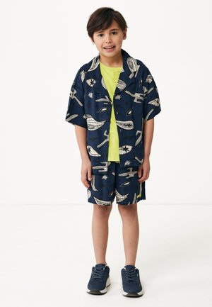 Kortærmet marineblå skjorte og matchende shorts med en kontrasterende hvidt print. Underneath en lys limegrøn T-shirt. Sorte sneakers fuldender outfittet.