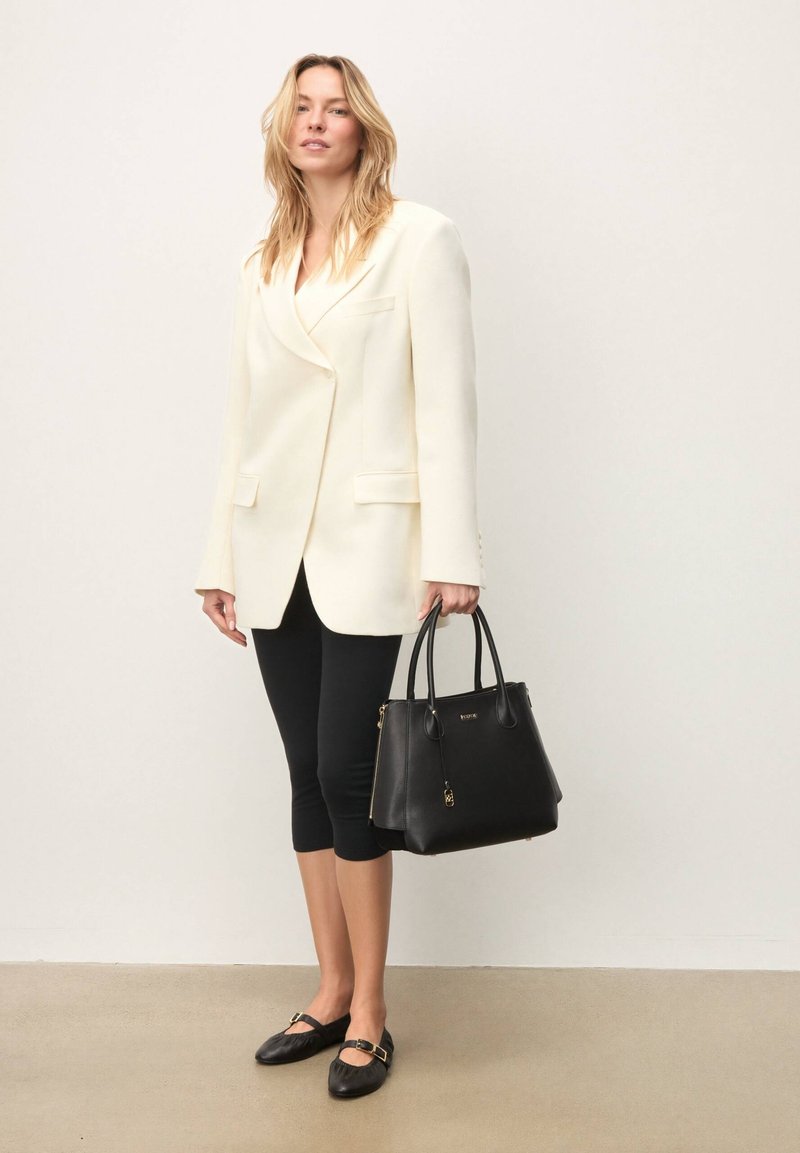 Sac à main en cuir noir avec des accents dorés, associé à un blazer blanc cassé et des leggings capri noirs. Le modèle porte des chaussures plates avec une boucle.