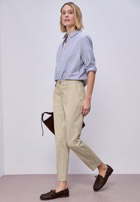 Kvinde iført blåt og hvidt stribet skjorte, beige bukser og brune loafers, holder en mørkebrun håndtaske, stående mod en ensfarvet væg.