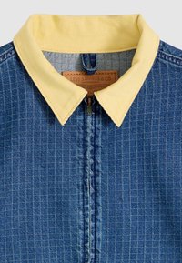 Τζιν μπουφάν με μικρό καρό μοτίβο, γιακά από κίτρινο κοτλέ ύφασμα, εμπρόσθιο φερμουάρ και ορατή δερμάτινη ετικέτα της μάρκας Levi Strauss & Co. στο εσωτερικό του γιακά.