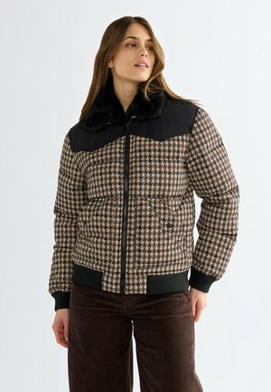 Frau trägt eine braune und schwarze Steppjacke mit Hahnentrittmuster, schwarzem Pelzkragen und schwarzen, gerippten Bündchen und Saum.