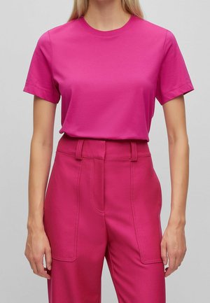 Vrouw draagt een felroze T-shirt met korte mouwen, ingestopt in hooggetailleerde, wijde roze broek met zichtbare voorzakken.