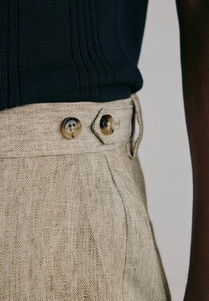 Lähikuva beigenvärisistä kalanruotokuvioisista housuista, joissa on kaksi sarvinappia ja kangasvyönlenkki, yhdistettynä tummaan neulepaitaan.