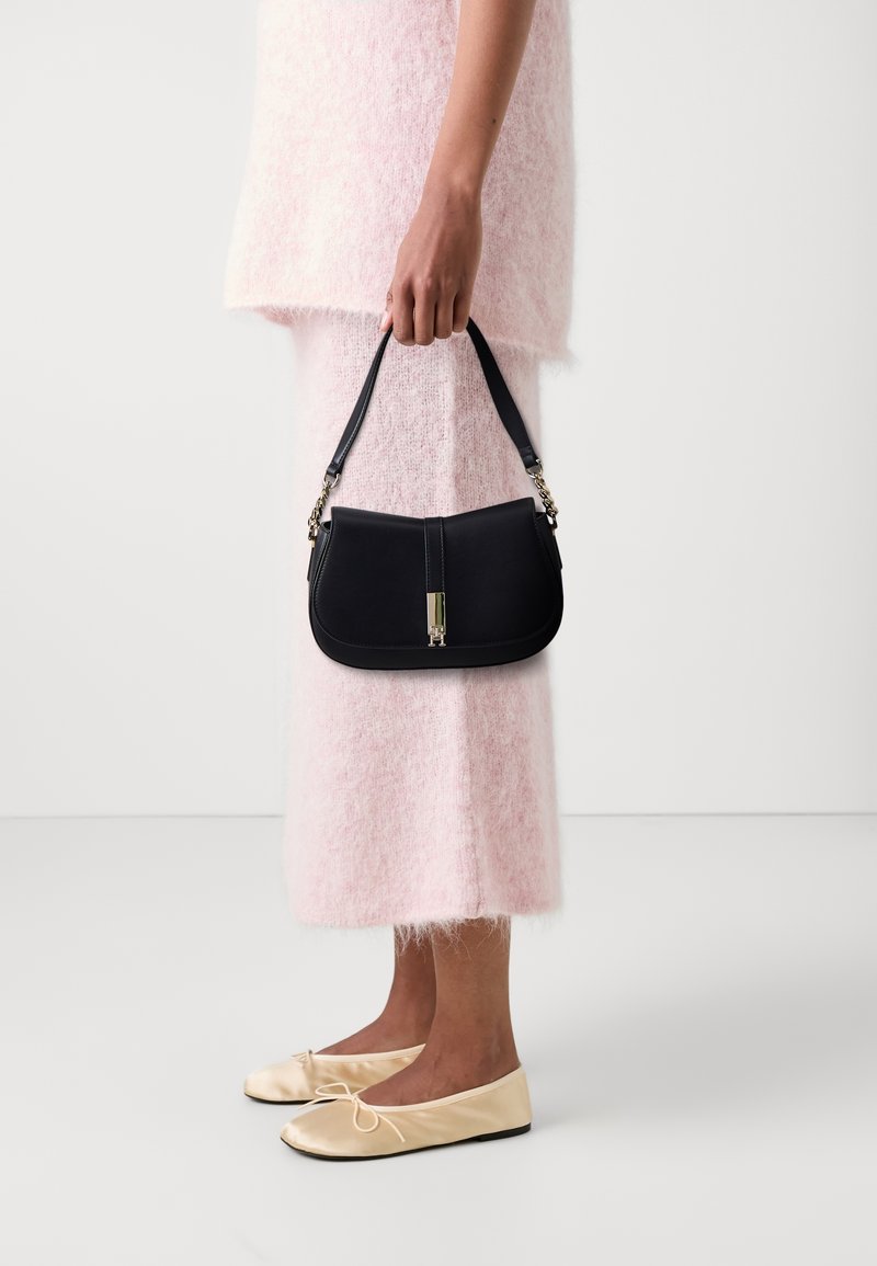 Personne portant une jupe duveteuse rose clair et un top, tenant un sac à main noir avec une fermeture dorée, chaussée de ballerines crème, debout sur un sol blanc.