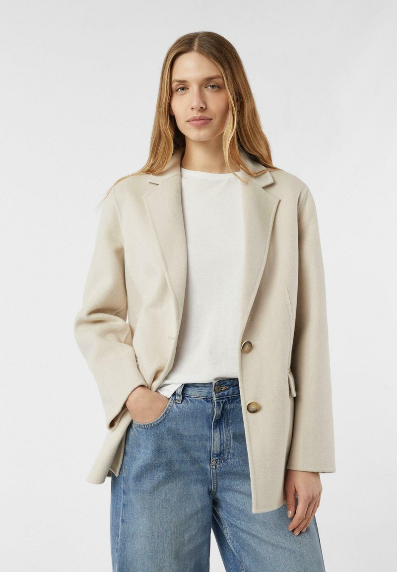 Cremefarbener Blazer mit leicht übergroßer Passform, ausgestattet mit gekerbten Revers, zwei großen Knöpfen und Seitentaschen, getragen über einem weißen Hemd.