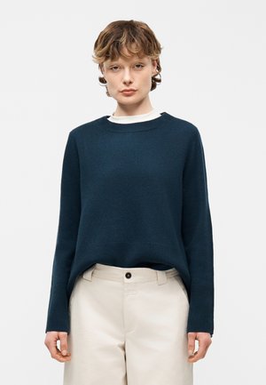 Young person with short wavy hair wearing a dark blue sweater over a white shirt and beige high-waisted pants standing against a white background.