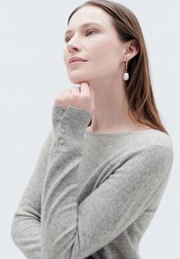 Maglione grigio a maglia con maniche lunghe e tre bottoni ai polsini. Il modello indossa un orecchino a goccia di perla; sfondo neutro.