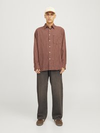 Chemise à manches longues à carreaux rouges et bruns avec une poche avant, assortie à un pantalon gris foncé ample et des chaussures en daim marron, accentuée par une casquette beige.