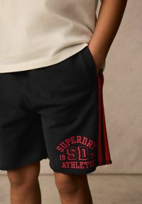 Schwarze sportliche Shorts mit roten Seitenstreifen, ausgefranstem Saum und einem großen roten Grafikaufdruck "SUPERDRY SD 1971" auf der Vorderseite. Baumwollmischgewebe.