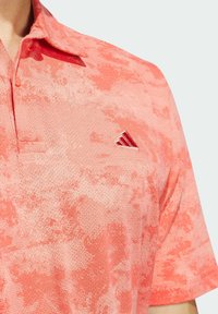 Lyhythihainen poolopaita teksturoidusta korallinvärisestä kankaasta, jossa on hillitty kuvio. Mukana on kolmen napin kiinnitys ja pieni punainen logo rinnassa.