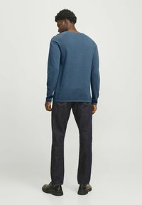 Blå, langærmet pullovertrøje med struktureret strik, båret over mørke jeans og sorte sko, set fra bagfra.