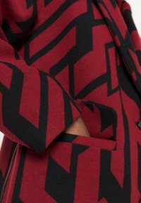 Rote und schwarz gemusterte Jacke mit geometrischen Formen, einer Seitentasche und strukturiertem Stoff mit einem maßgeschneiderten Ärmel.