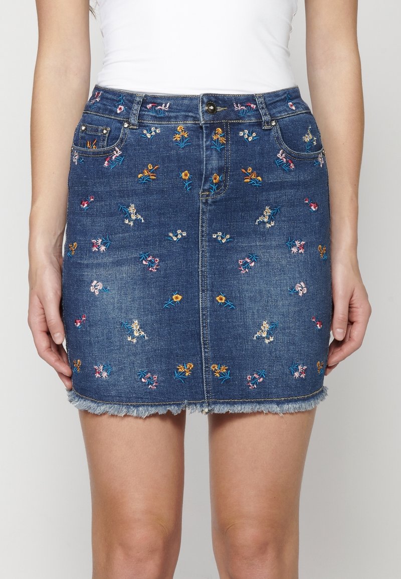 Jupe courte en denim avec petite broderie florale multicolore, fermeture à bouton et zip à l’avant, et ourlet effiloché portée avec un haut blanc.