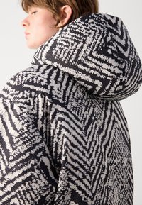 Giacca con cappuccio dal motivo tessuto testurizzato in bianco e nero a zigzag. Vestibilità oversize, che mette in mostra i dettagli e il design del tessuto.