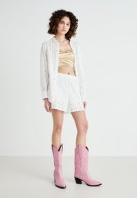 Chemise blanche légère avec des accents floraux, crop top en satin doré, shorts blancs avec une bordure en dentelle, et bottes de cowboy roses avec un talon noir.