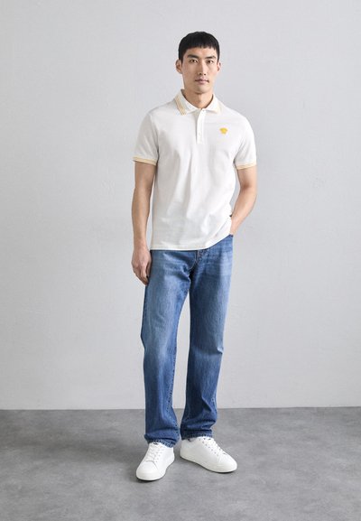 Tricou polo alb cu accente galbene, purtat cu blugi albaștri și teniși albi. Design simplu, mâneci scurte și guler.