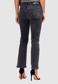 Femme portant un jean bootcut noir et des sandales à talons hauts noires, montrée de dos sur fond uni.