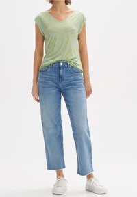 Žena v světle zeleném tričku s výstřihem do V, krátkým rukávem, zasunutém do vysokých modrých džínů s kratší nohavicí a bílých teniskách na bílém pozadí.