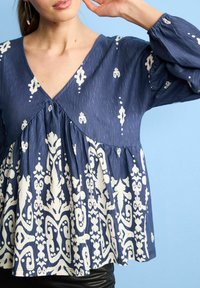 Blauer, gemusterter Blouson-Oberteil mit V-Ausschnitt, das weiße Blumen- und abstrakte Designs, strukturierten Stoff und geraffte Details unterhalb der Brust aufweist.