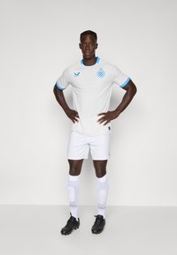 Balta sportinė marškiniai su mėlynais akcentais ir logo, derinami su baltais šortais ir raštuotomis kojinėmis. Juodi futbolo batai su tekstūruotu dizainu.