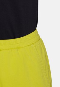 Czarny top wsunięty w spodnie z neonowożółtym elastycznym pasem i widocznym przeszyciem kieszeni bocznych na jednolitym tle.