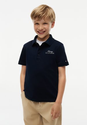 Uśmiechnięty młody chłopiec o blond włosach, ubrany w granatową koszulkę polo Tommy Hilfiger i beżowe spodnie, stojący z rękami w kieszeniach.