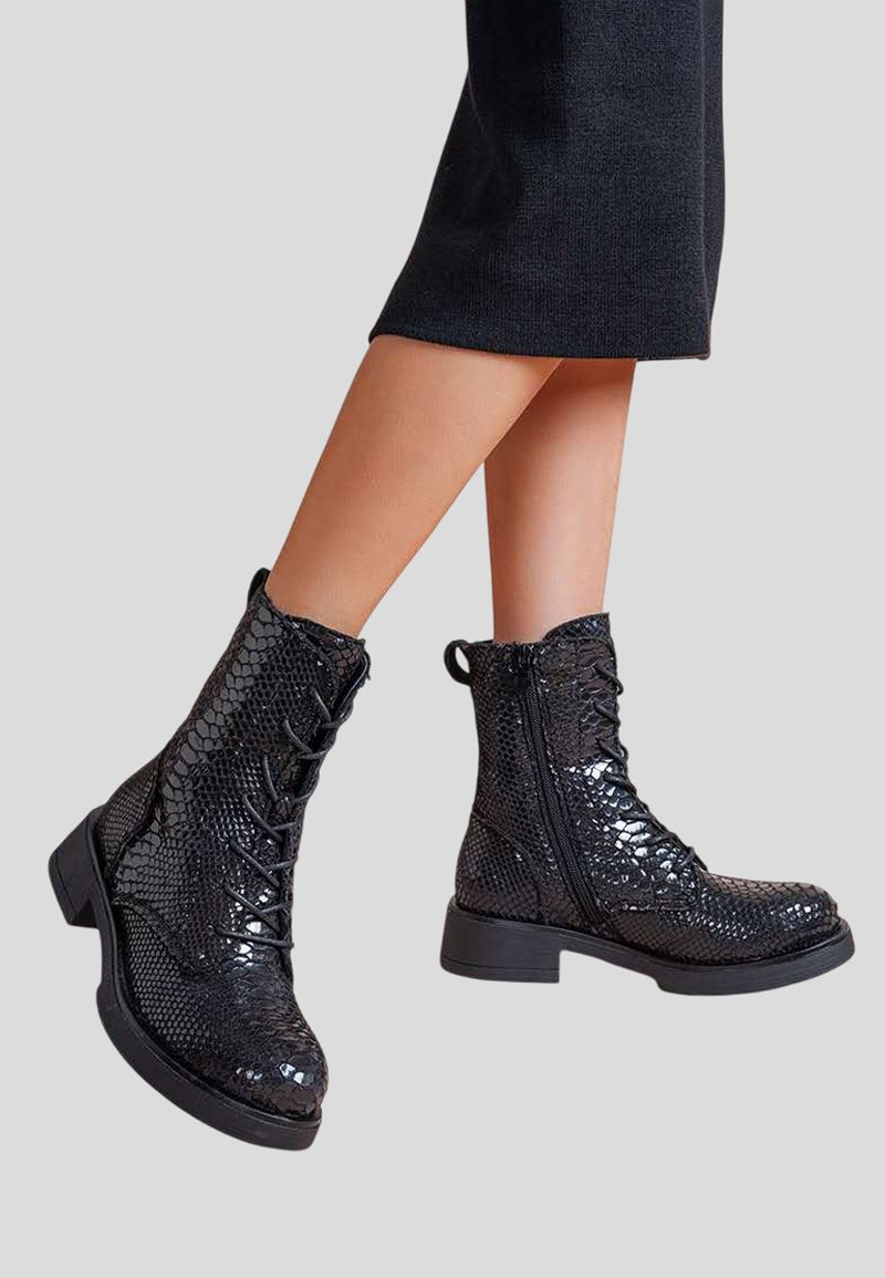 Bottines noires brillantes avec texture en cuir de serpent, lacets à l'avant, fermetures éclair sur le côté, portées avec une jupe noire sur un fond clair.