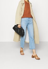 Beige lange jas over een oranje gestructureerde trui, lichtblauwe cropped jeans en bruine haksandalen. Zwarte gestructureerde clutch tas in de hand.