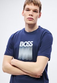 Temno modra bombažna majica s potiskanim napisom "BOSS" in valovitim gradientnim vzorcem v sivih odtenkih. Klasičen okrogel izrez.